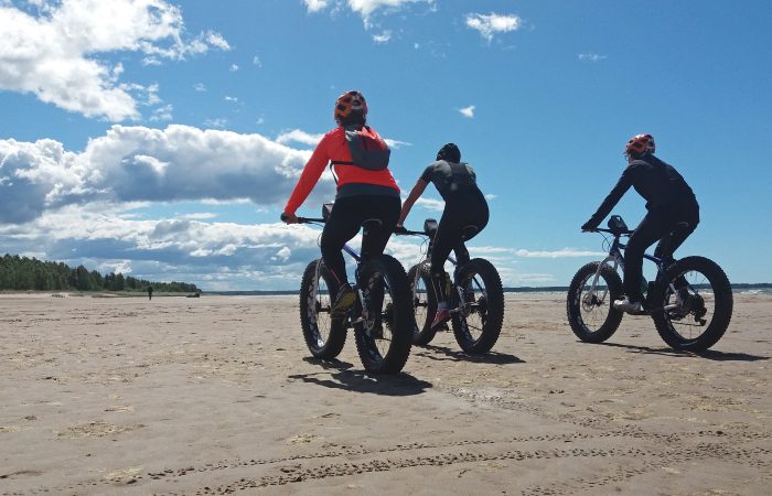 Kolme pyöräilijää hiekkarannalla.