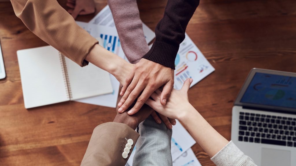 Hands of six people on top of each other, a table with papers and a laptop on the background.