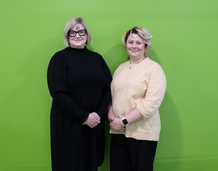 Two women standing in front of a green wall.