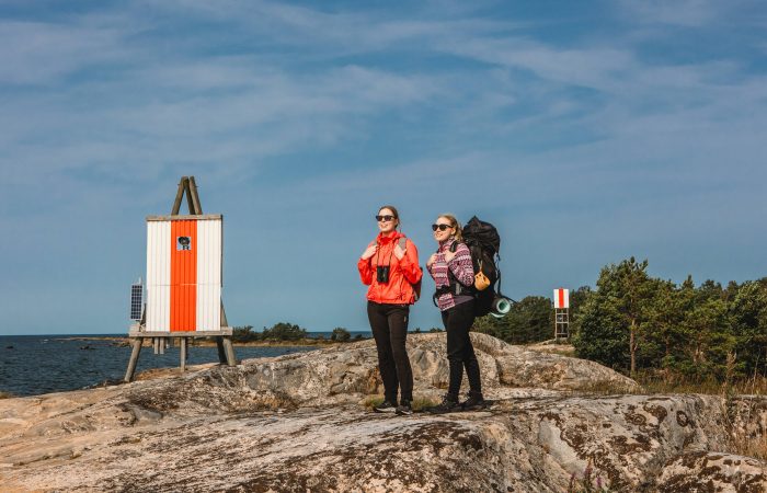 Kaksi naista rinkat selässä kallion päällä meren rannalla.