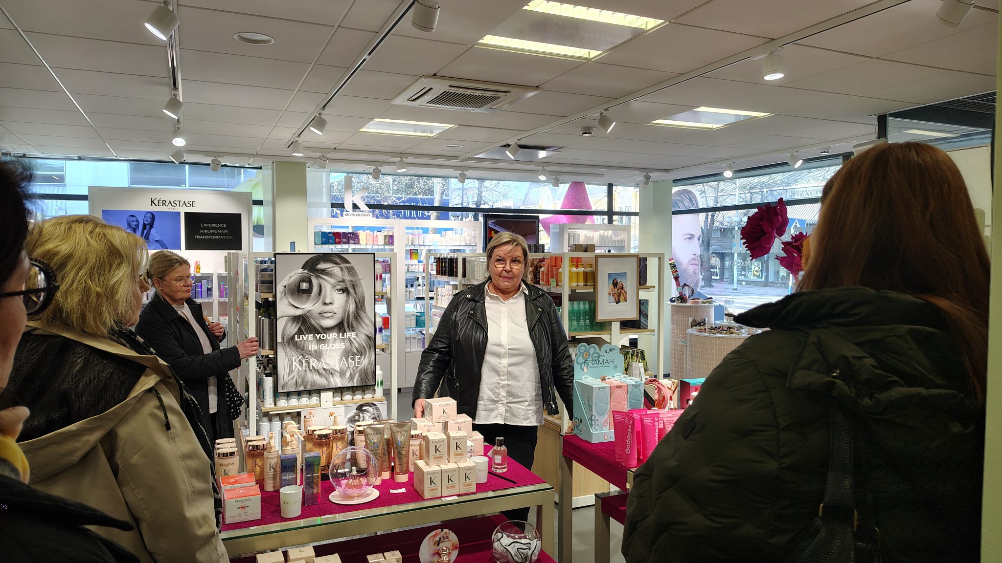 In a cosmetics store, a specialist is presenting products to a small group. In the foreground, shelves display skincare and beauty products along with large product images.