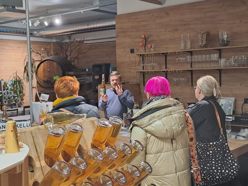 Pieni ryhmä asiakkaita kuuntelemassa esittelijää, joka pitää kädessään juomapulloa siiderituotantotilan maistatuspisteessä. Taustalla on puisia hyllyjä ja suuria tynnyreitä.