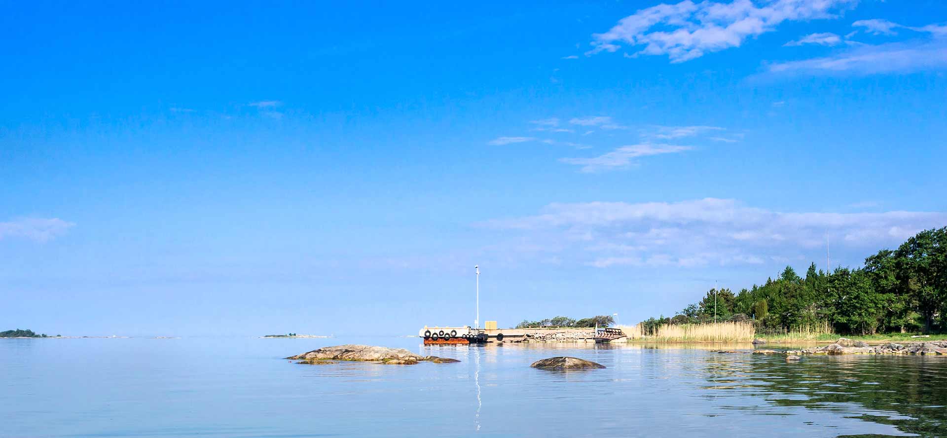 Kapea vesiuoma avautuu merelle, reunoilla ruovikkoa ja kallioita, taustalla sininen taivas ja rauhallinen saaristomaisema.