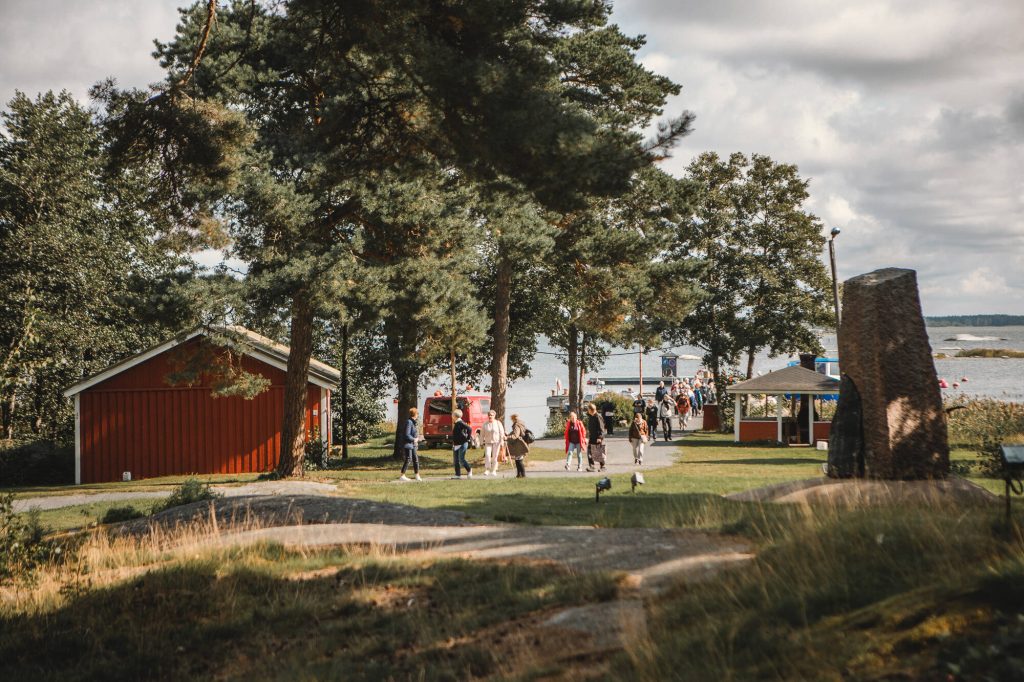 Kuvituskuva. rakennuksia, järvimaisemaa ja ihmisiä.