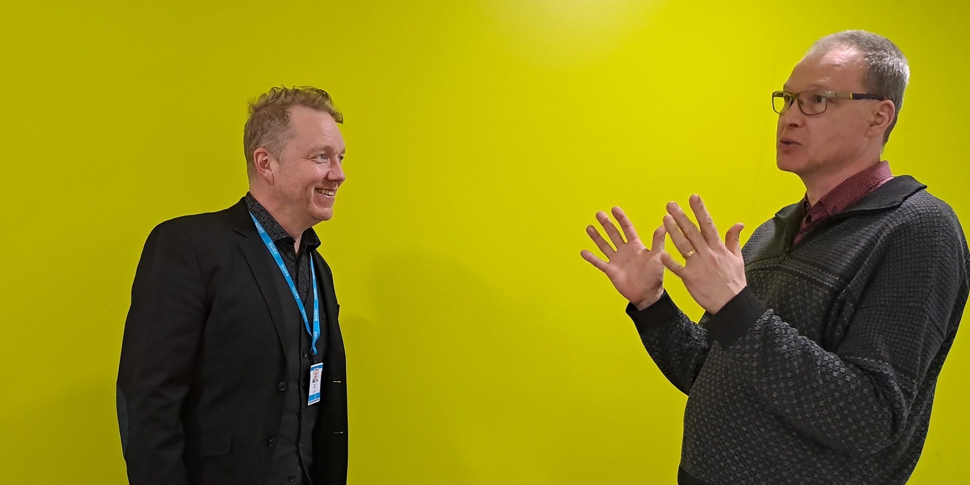 Kaksi miestä keltaisen taustan edessä keskustelemassa toisilleen.