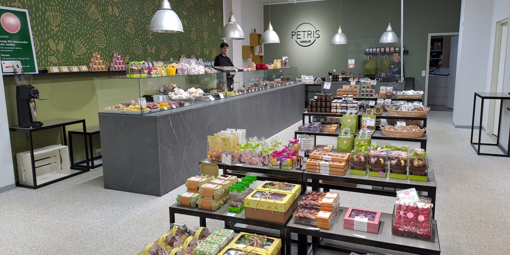 A candy shop with long display tables and multiple tiers filled with colorful chocolate and caramel products. In the background, a shop assistant stands behind the counter in a green‑toned store.
