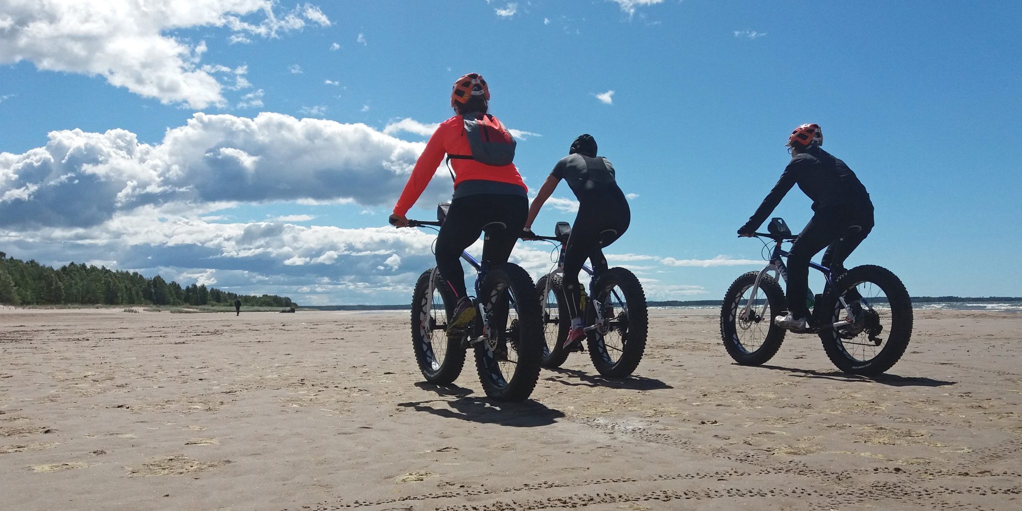 Kolme pyöräilijää hiekkarannalla.