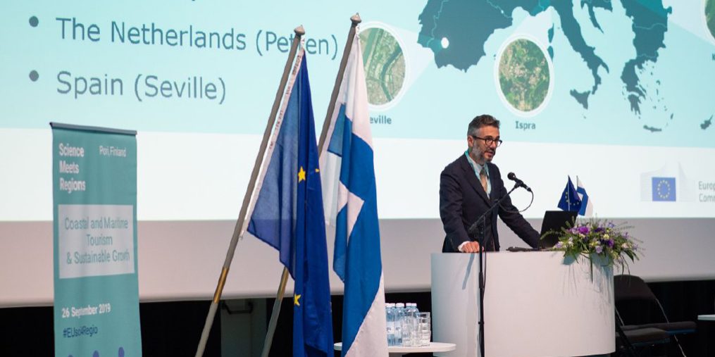 Henkilö pitämässä puhetta puhujakorokkeella Science Meets Regions -tapahtumassa. Taustalla Euroopan karttakuva.