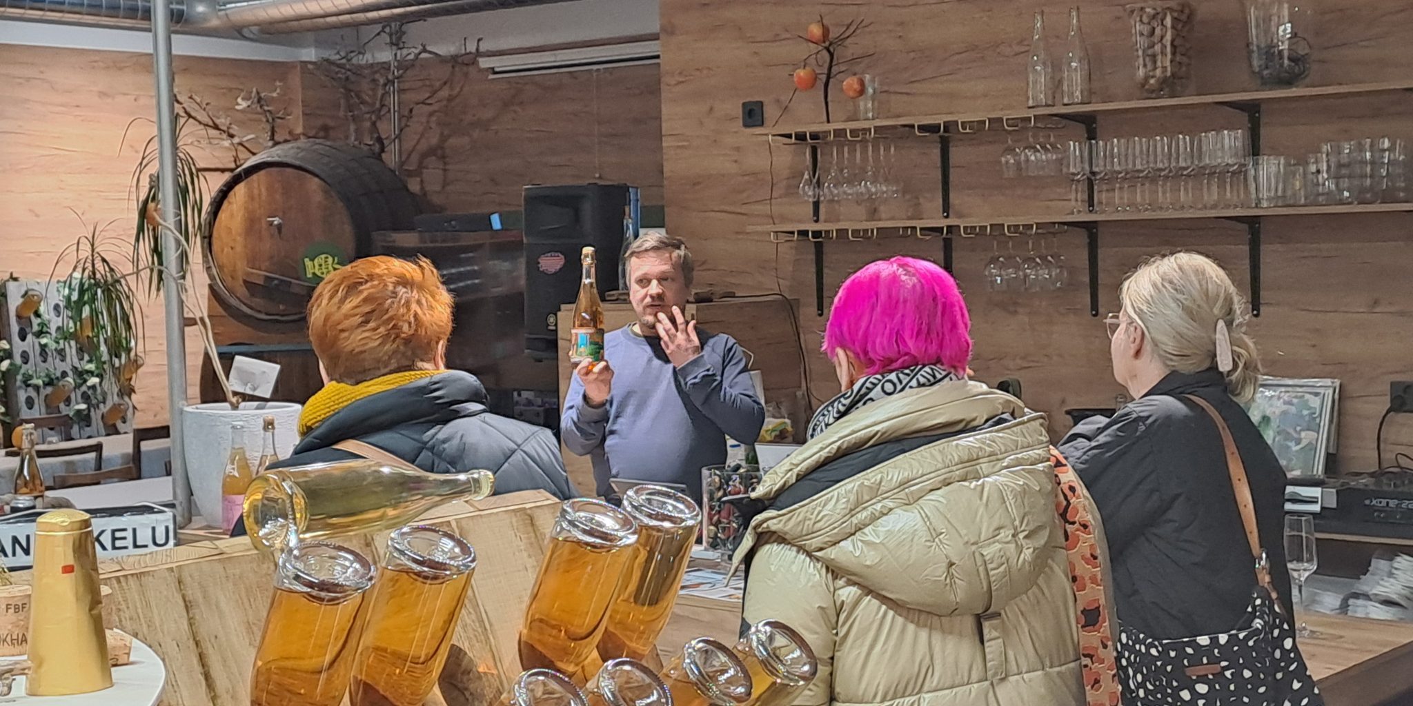 A small group of customers listening to a presenter who is holding a drink bottle at a tasting station in a cider production facility. In the background, there are wooden shelves and large barrels.