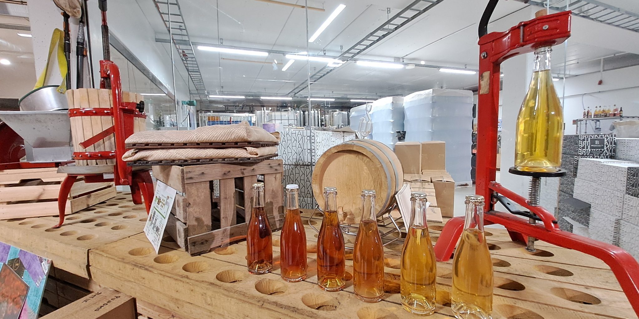 On a table, different shades of drink bottles are lined up in front of a cider production area. In the background, barrels, production equipment, and storage space can be seen.