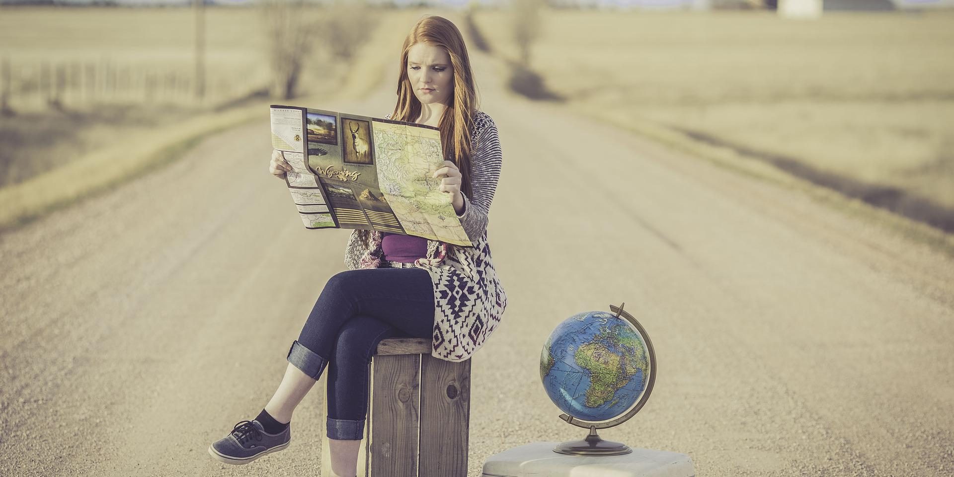 Young woman sitting on a suitcase in a middle of a road, a map in her hands.