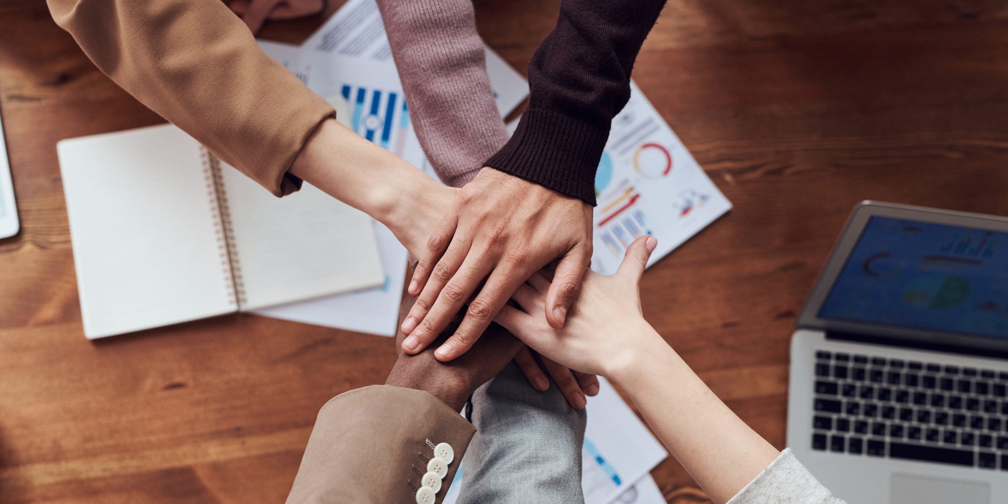 Hands of six people on top of each other, a table with papers and a laptop on the background.